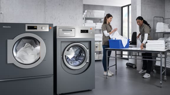 Two people work in a bright laundry room with industrial machines, folding towels and handling laundry supplies.