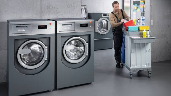 An employee with a cleaning trolley enters a room with grey washing machine and tumble dryer.