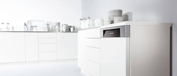 Range of Miele Professional Tank dishwasher: One throughput dishwasher and three tank dishwasher in a row, opened door equipped with baskets, glass and cutlery on grey background. In front another tank dishwasher with opened door and equipped.