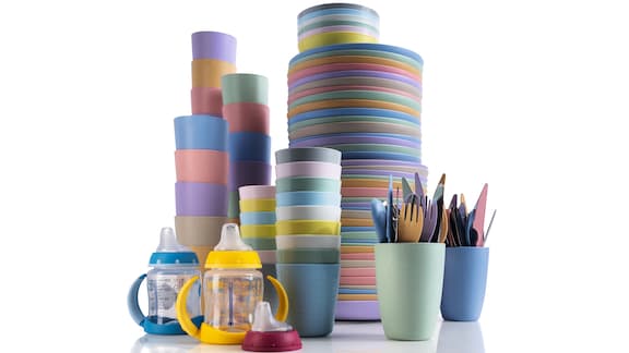Stacked colourful reusable plastic plates and cups on a white background. In front, colourful cups with reusable cutlery in it and two drink-training-cups for children.  