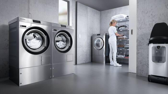 Dos lavadoras Benchmark Performance Plus están una al lado de la otra en la lavandería. Al fondo, un cuidador vestido de blanco junto a una secadora Benchmark Performance Plus clasifica la ropa en un estante.