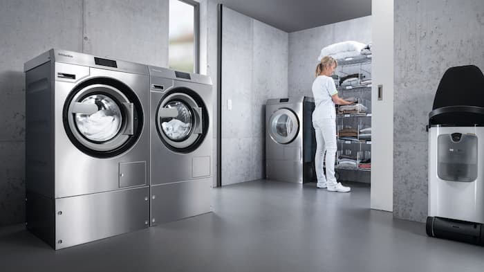 Kaksi Benchmark Performance Plus ‑sarjan pyykinpesukonetta vierekkäin pesutuvassa. Taustalla valkoisiin pukeutunut hoitaja seisomassa Benchmark Performance Plus ‑kuivausrummun vieressä lajittelemassa pyykkiä telineeseen.