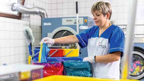 Εργαζόμενο άτομο στην TGS Teegen τακτοποιεί τα σύνεργα καθαρισμού σε τροχήλατο για είδη καθαρισμού. Στο φόντο διακρίνονται συσκευές Miele Professional.