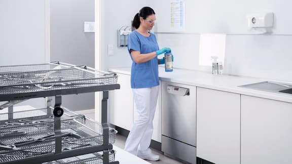 Βοηθός ιατρού απεικονίζεται να τακτοποιεί το δωμάτιο συσκευών επανεπεξεργασίας στο ιατρείο. Σε πρώτο πλάνο διακρίνεται ένα δοχείο για τα χημικά υλικά καθαρισμού ProCare Med.