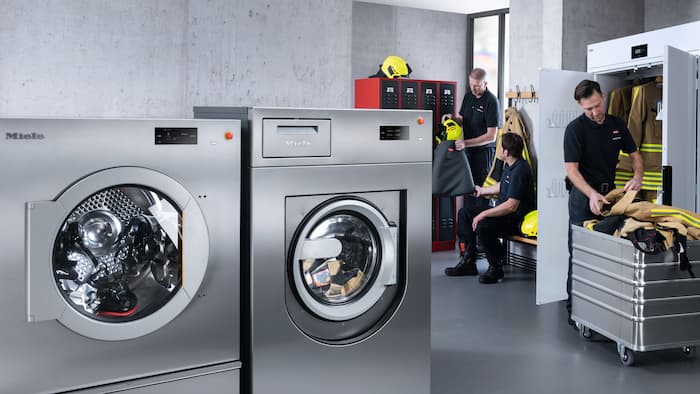 Three firefighters stow their personal protective equipment before it is washed or dried. The room also contains the Miele Benchmark Performance Plus washing machine and the Nimo drying cabinet.