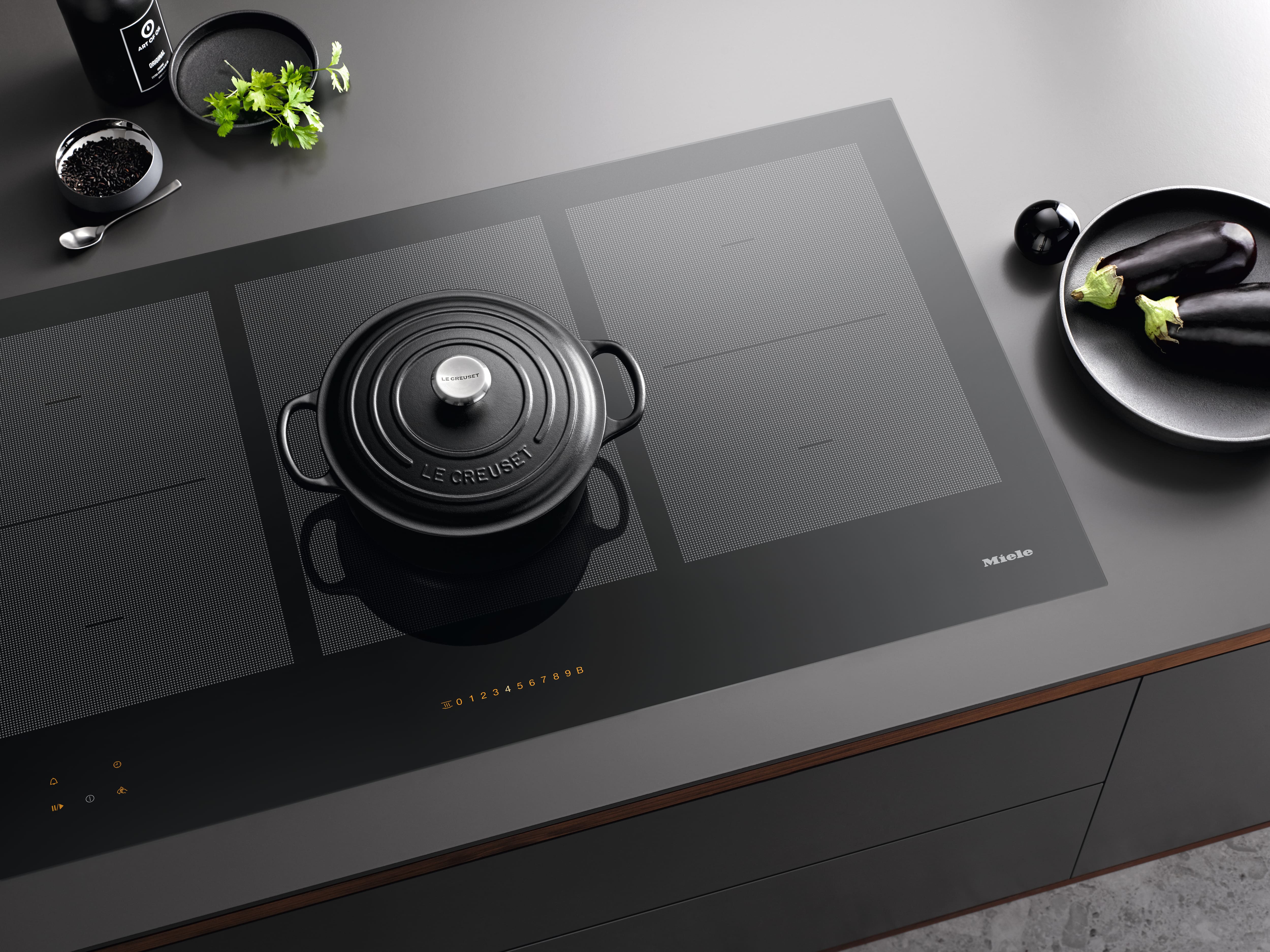 Parrilla de inducción Miele con una olla de hierro fundido, rodeada de hierbas frescas y berenjenas sobre una encimera de cocina minimalista.