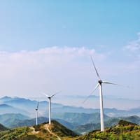 Kolme tuulivoimalaa vihreiden vuorten huipuilla, taustalla sininen taivas