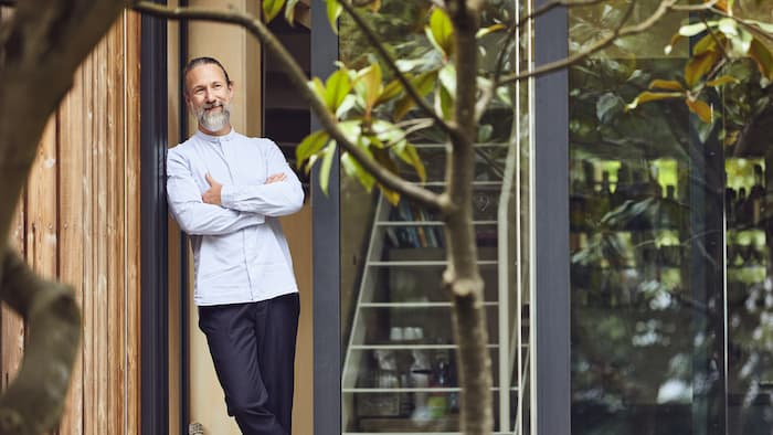 A man with a white shirt and beard leans against a wooden house wall. He smiles and looks into the distance.