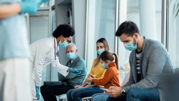 Várias pessoas estão sentadas numa sala de espera de um médico com máscara facial.