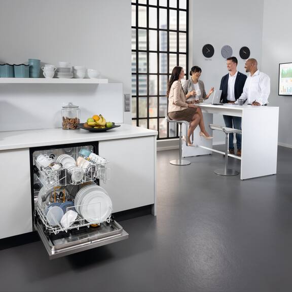 Miele Professional MasterLine oppvaskmaskin som er åpnet og fylt med servise, står i et kontorkjøkken mens et møte pågår i bakgrunnen