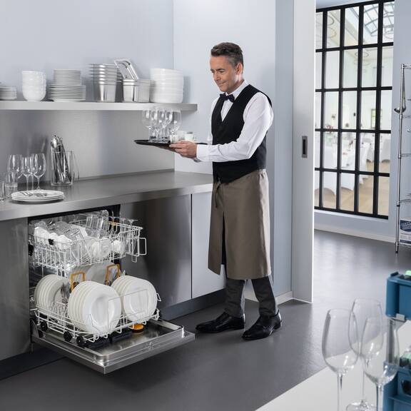 Waiter puts glasses into opened Miele Professional MasterLine dishwasher loaded with dishes in a restaurant kitchen