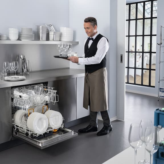 Een kelner zet glazen in een geopende en met serviesgoed beladen Miele Professional MasterLine-vaatwasser in een restaurantkeuken