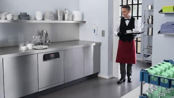 Exemple illustré d’un restaurateur apportant de la vaisselle sale dans la cuisine où se trouve un lave-vaisselle Miele