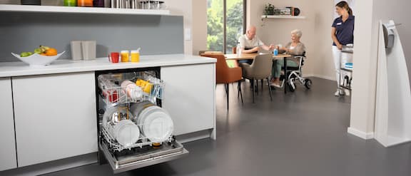 Kitchenette in a care and nursing home with opened and equipped dishwasher. Decoration and tea pots on a shelf above the kitchenette. A nurse is pushing a service cart in the background. In the background, two residents play cards.