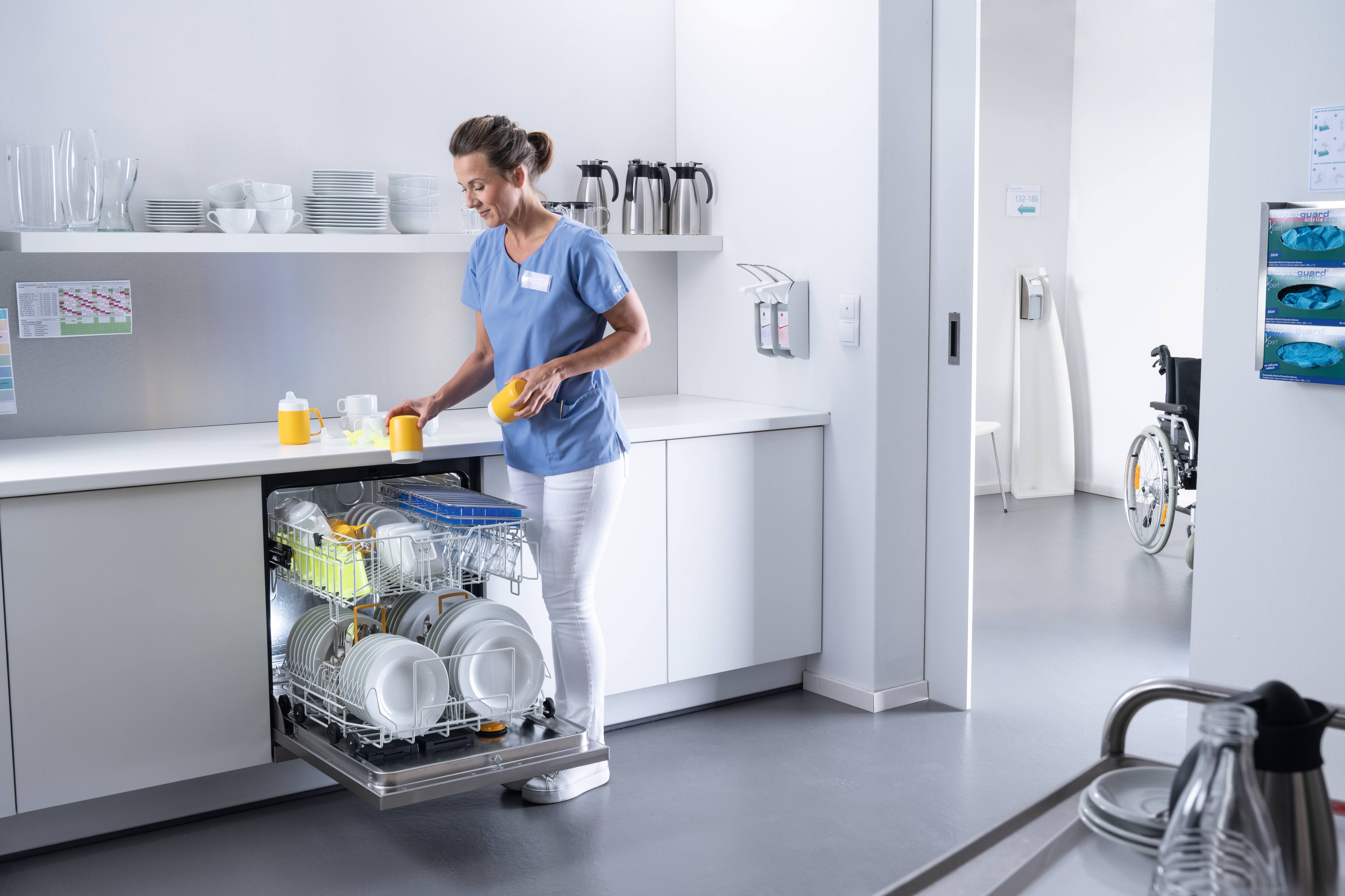 Pečovatelka uklízí v kuchyni hrnky do otevřené myčky Miele Professional MasterLine naplněné nádobím