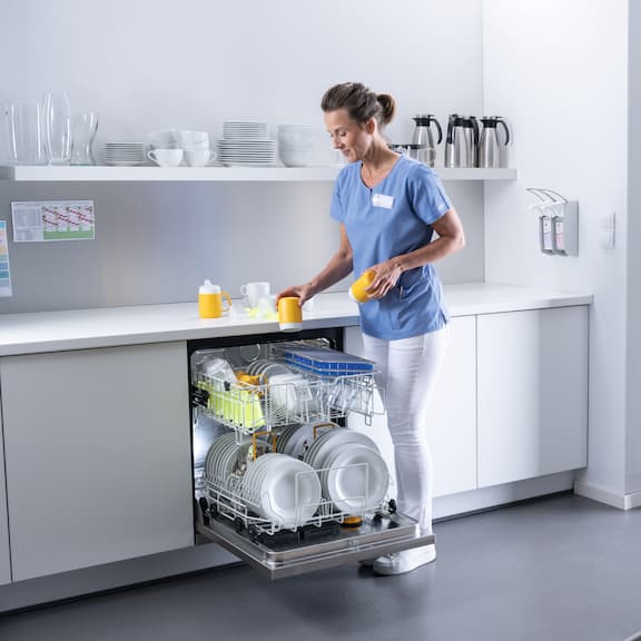 Caregiver puts cups into opened Miele Professional MasterLine dishwasher loaded with dishes in a kitchen