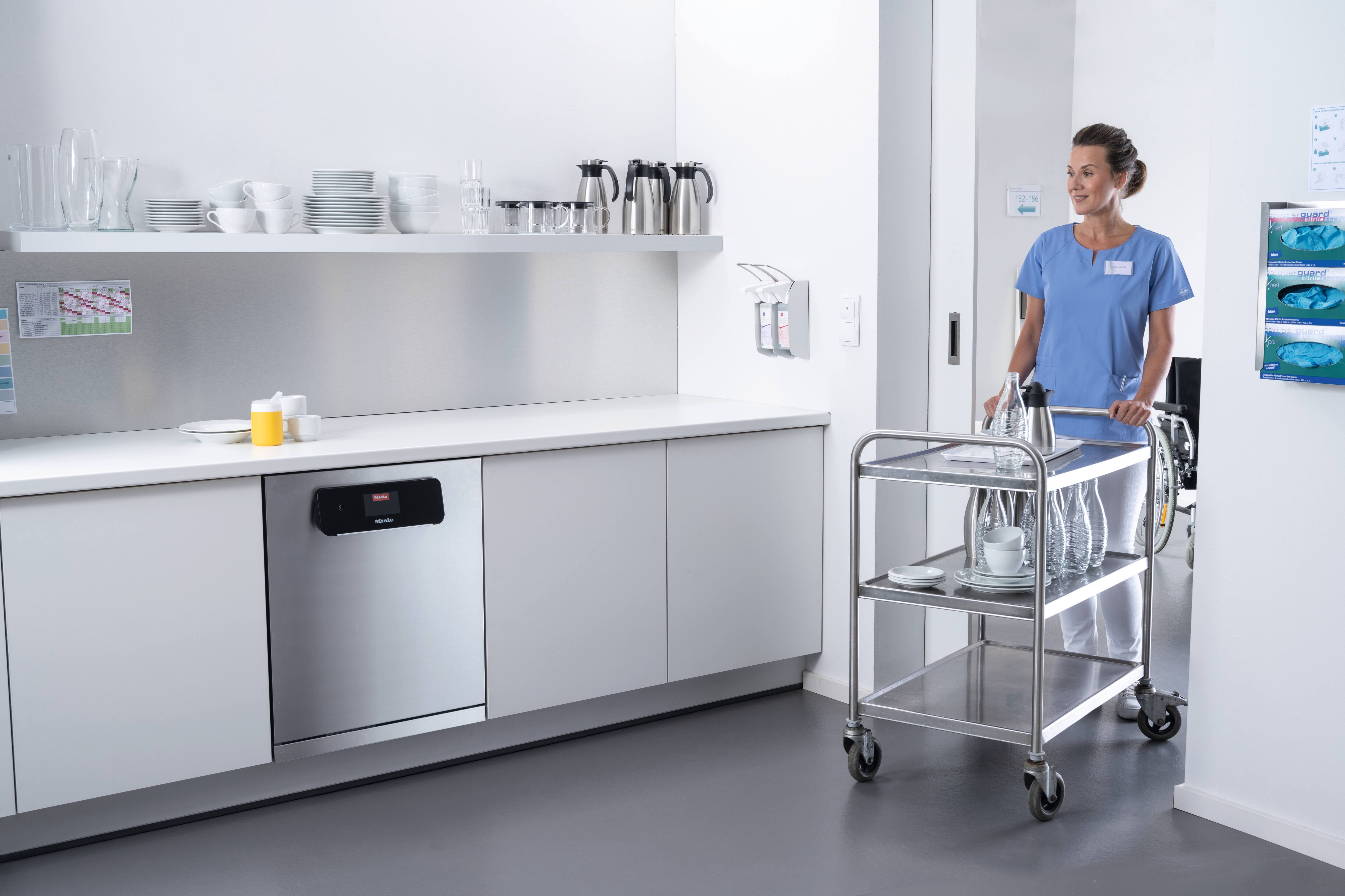 Modern tvättanläggning med Miele Professional Benchmark-tvättmaskin i förgrunden. Två medarbetare i grå arbetskläder viker och sorterar nytvättade handdukar. 