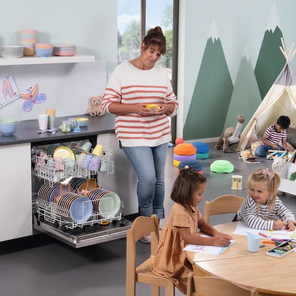Educadora ao lado de uma máquina de lavar louça MasterLine da Miele Professional aberta e carregada com louça num infantário com crianças a brincar em primeiro plano
