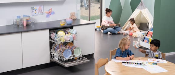 Kitchenette in a nursery with opened and equipped dishwasher. Next to it a nurse. Some plastic crockery on the worktop. In foreground two children drawing pictures at a table. In the background a tent with a nurse playing with a little girl.