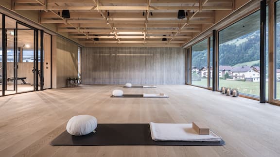 Een ruime kamer met een houten vloer, waarin verschillende yogamatten met kussens en handdoeken liggen.  