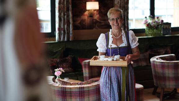 A women in a dirndl, carrying a tray with white crockery, smiling.