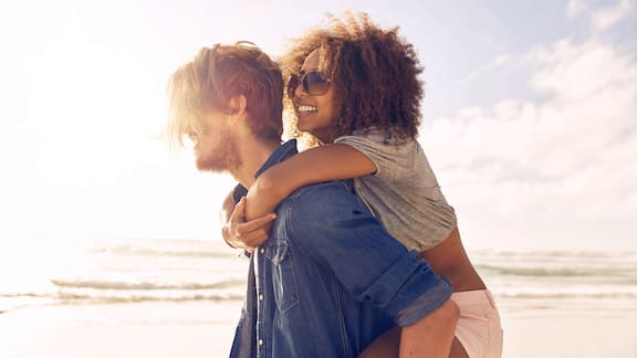 Een man draagt een vrouw op de rug, op de achtergrond is het strand te zien 