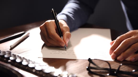 Person writing with a pen on a sheet of paper. A pair of glasses on the table.   
