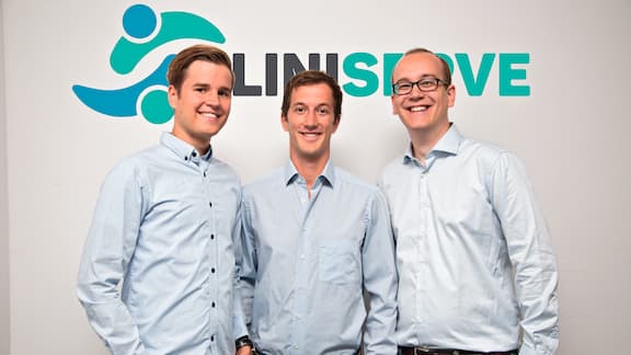 Die Cliniserve-Gründer Jaakko Nurkka, Julian Nast-Kolb und Quirin Körner stehen nebeneinander vor einer weißen Wand bedruckt mit dem Cliniserve Logo.  