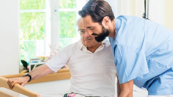 A young dark-haired care worker helps an elderly gentleman out of bed 