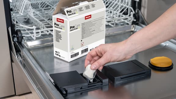 Porte de lave-vaisselle ouverte avec un paquet de tablettes pour lave-vaisselle. Une main insère une pastille dans une trappe ouverte pour le détergent.