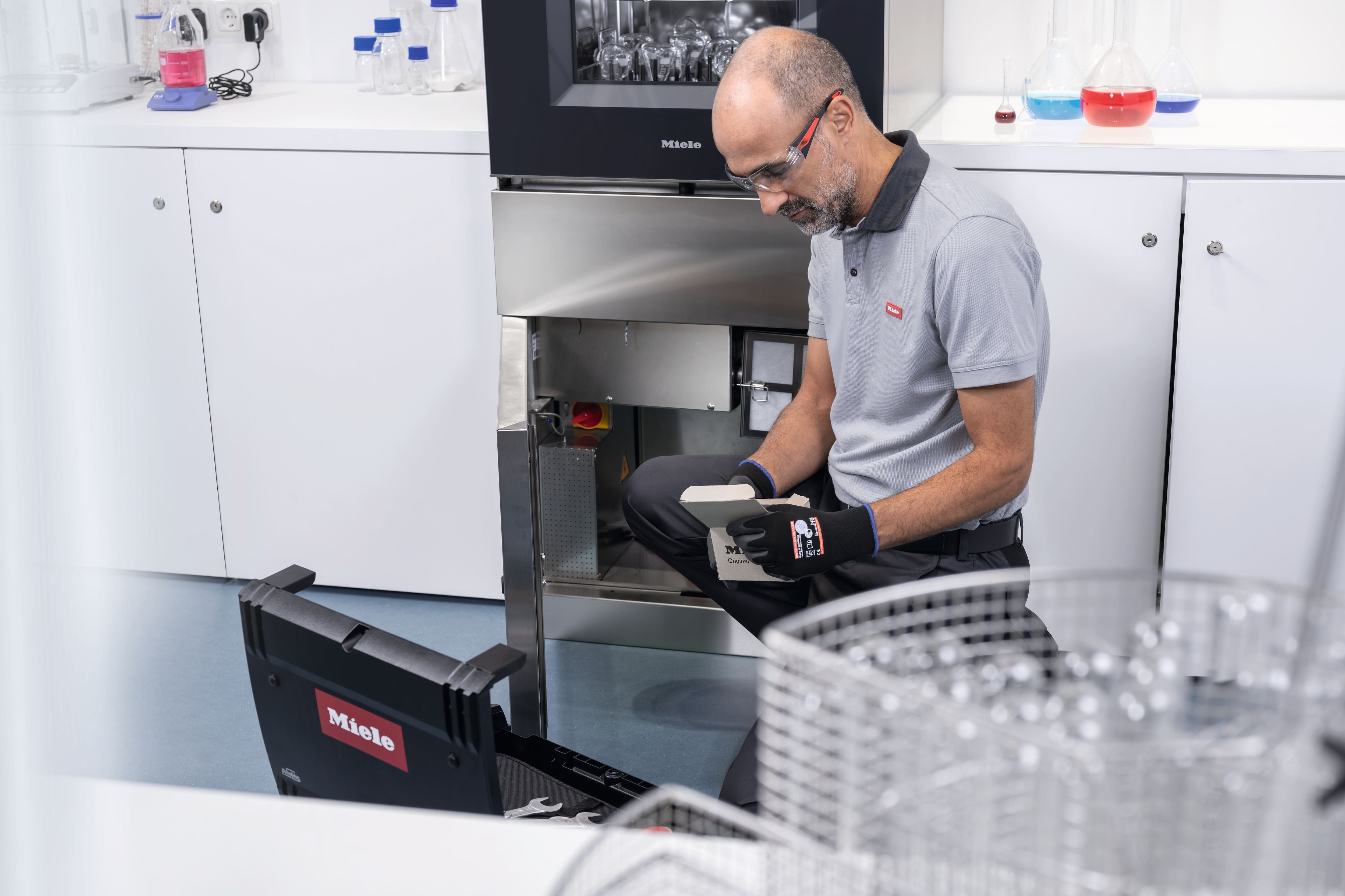 Ein Servicetechniker kniet vor einem SlimLine Laborspüler und hält ein Ersatzteilpaket in der Hand.