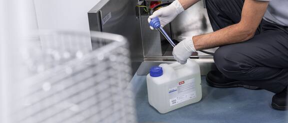 Un technicien de service est agenouillé devant un laveur de laboratoire SlimLine et insère une lance de dosage dans un bidon de produit ProCare Lab.
