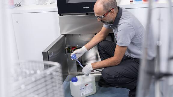 A service technician kneels in front of a laboratory washer and configures the automatic dosing of a cleaning agent.