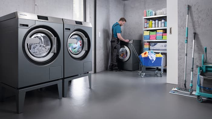 In een waskelder staan Benchmark Performance-wasmachines en -droogautomaten. Op de achtergrond bedient een schoonmaker de besturing van de Benchmark-droogautomaten.