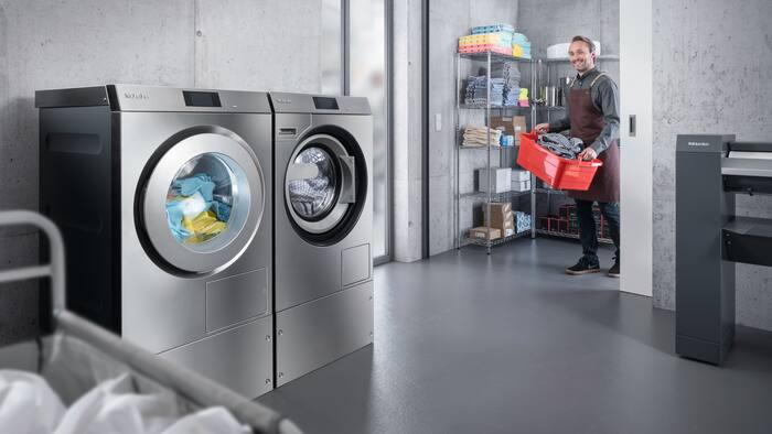 Un empleado de un restaurante con una cesta llena de paños de cocina entra en el cuarto de lavado de uso colectivo en el que hay una secadora y una lavadora de la serie Benchmark Performance Plus.