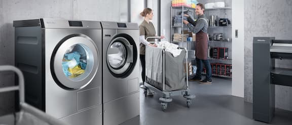 Bild aus einer Wäscherei mit einer Miele-Waschmaschine und einem Miele-Trockner sowie Personen, die Wäsche transportieren.