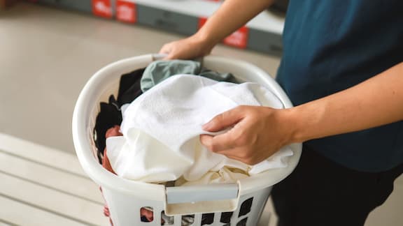 Ein weißer Wäschekorb voll mit Wäsche wird von einer Person in einem Waschsalon getragen. 