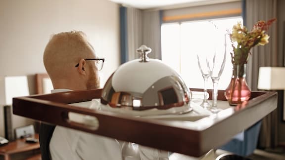 A hotel employee carries a tray with serving bell into a room 