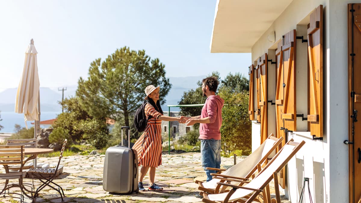 A traveller is welcomed by her host in front of a holiday home 