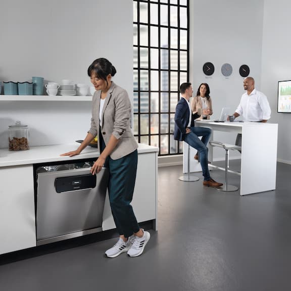 Two employees are standing in the office kitchen, one is sitting on a bar stool. A fourth employee is operating the Miele MasterLine dishwasher.