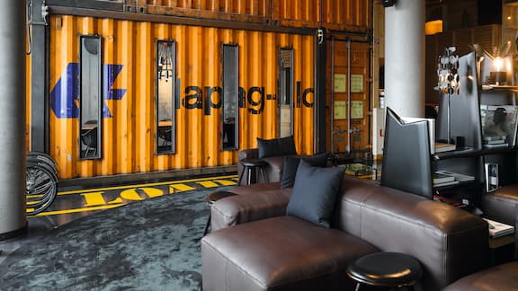 Lobby d‘hôtel avec un coin salon marron et un mur décoré d‘un véritable conteneur maritime Hapag-Lloyd. 