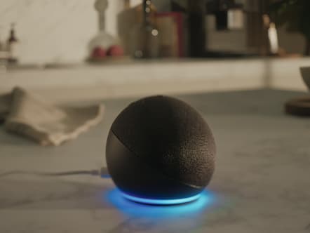 上部が布製で、青く光るライトリングが付いた球形のスマートスピーカーが、充電ケーブルが接続されたキッチンカウンターの上に置かれている。