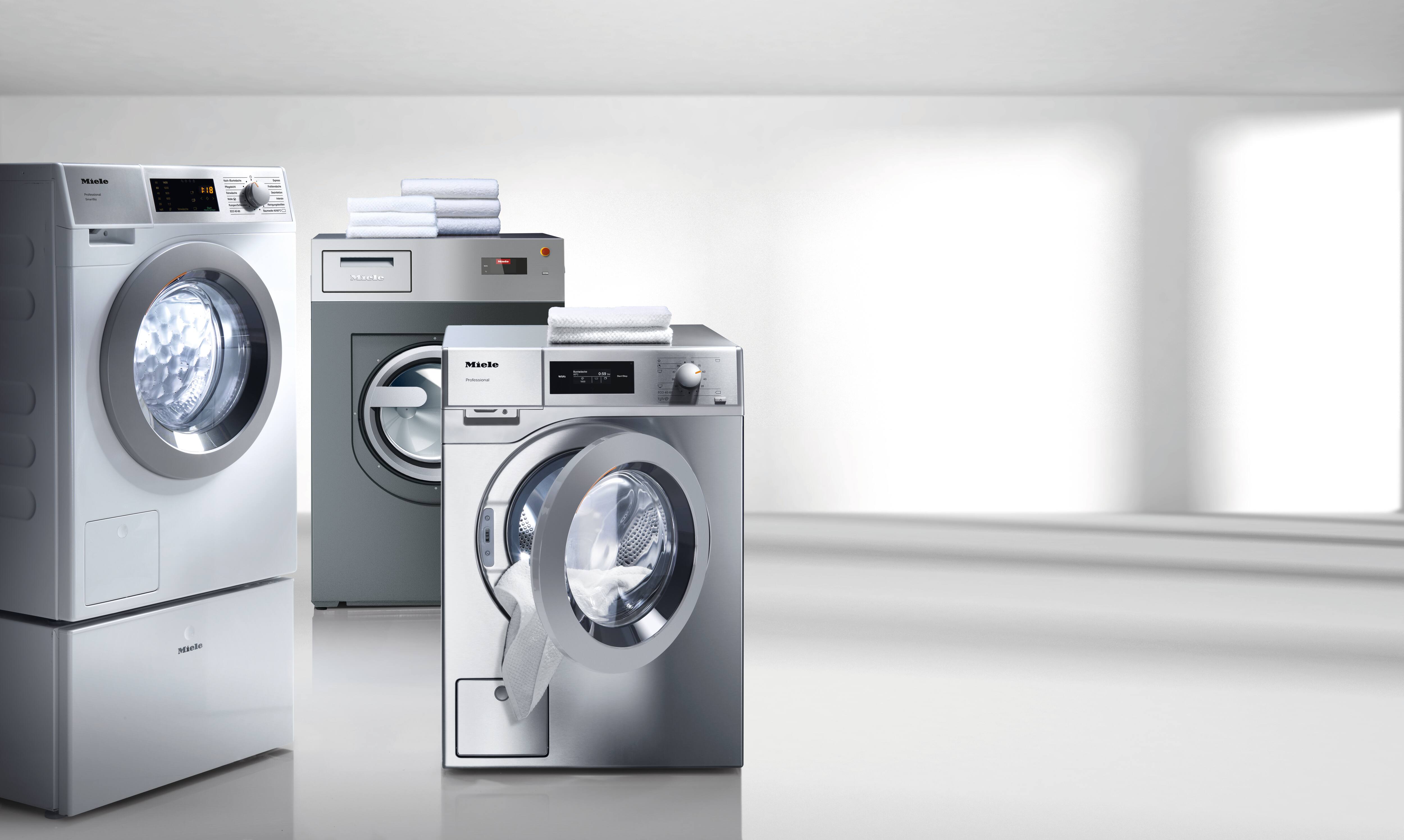 A room contains commercial washing machines from the Miele Professional SmartBiz, Little Giants and Benchmark series. Towels have been left on Little Giants and Benchmark washing machines. The door of the Little Giants washing machine is half open and a towel is sticking out.