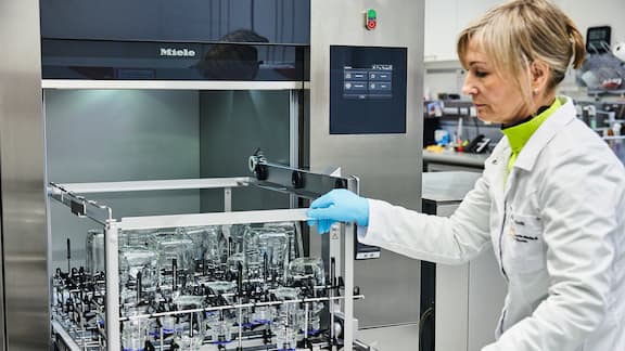 Eine Labormitarbeiterin schiebt einen mit Laborglas bestückten Korb in einen großen Laborspüler.