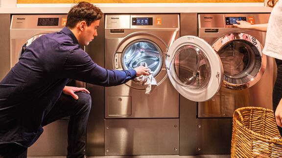 Ein junger Mann belädt eine Miele Professional Kleine Riesen Waschmaschine in einem schicken Waschsalon. 