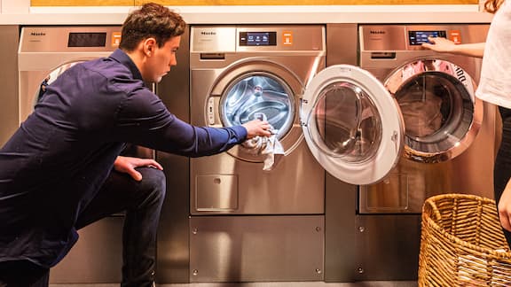 Ein junger Mann belädt eine Miele Professional Kleine Riesen Waschmaschine in einem schicken Waschsalon. 