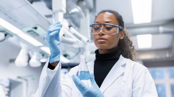 Eine Labormitarbeitern mit Schutzbrille und Laborkittel hält Laborglas in der Hand und schaut dies konzientriert an 