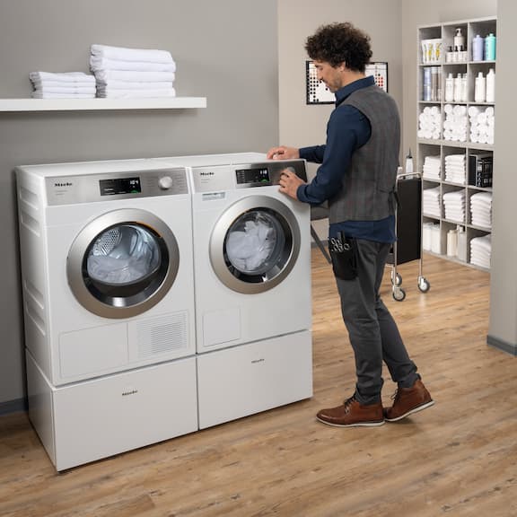 En frisör står vid en Miele SmartBiz-tvättmaskin PWM 1108 och ställer in ett tvättprogram. Bredvid PWM 1108 står Miele SmartBiz-värmepumpstumlare PDR 1108 HP