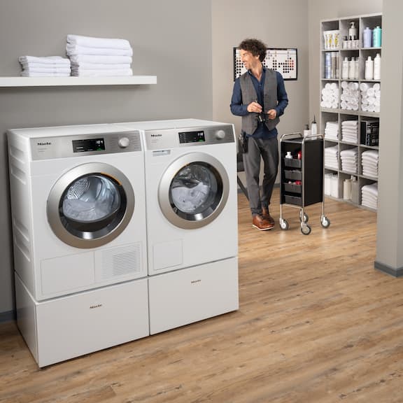 A hair dresser is working in a hair salon in the background. In the forefront the Miele SmartBiz washing machine and dryer are standing next to each other in the laundry room.