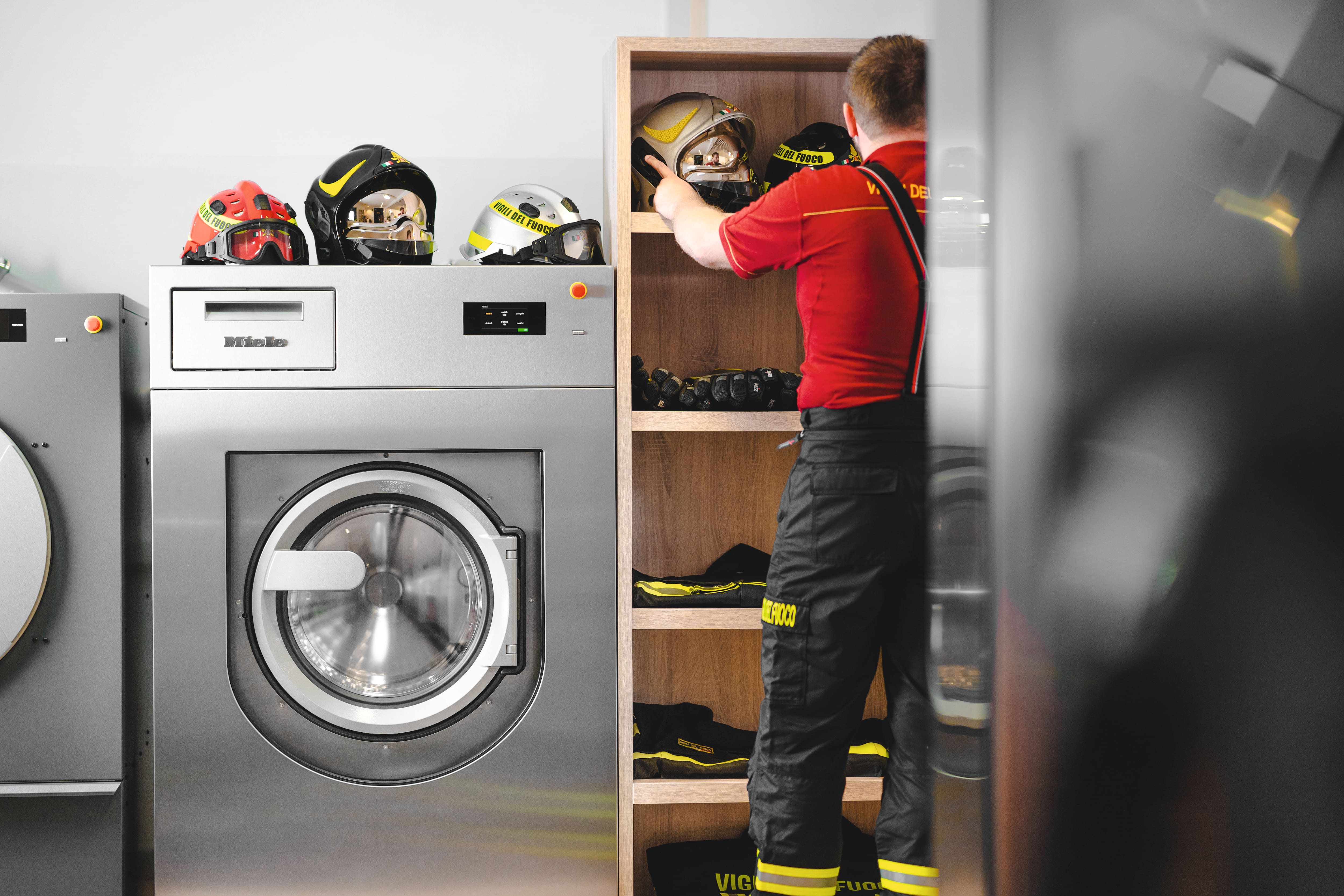Ein Feuerwehr- mann lagert verschiedene Helme in einem Schrank ein. Einige Helme liegen ebenfalls auf der Miele Benchmark Performance Plus Waschmaschine ab, welche neben ihm steht.